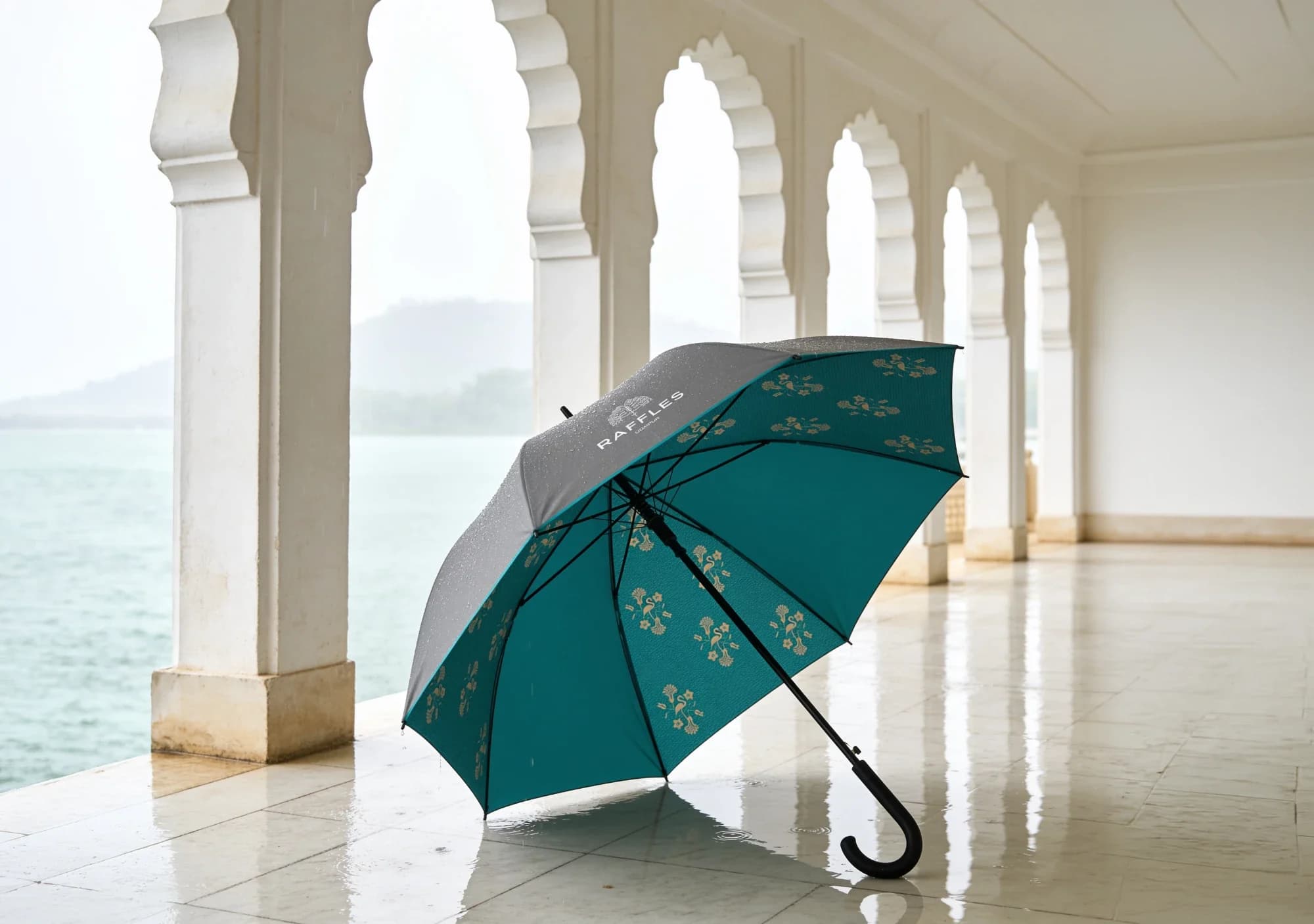 The Raffles Udaipur umbrella, open on a wet marble floor in an arcade of scalloped arches beside the lake